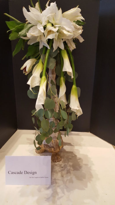 A vertically shaped plant with a grouping of white calla lilies at the top, and then 6-8 white calla lilies hang upside down from under the grouping at top falling down into vertical strands of Aspen vines.